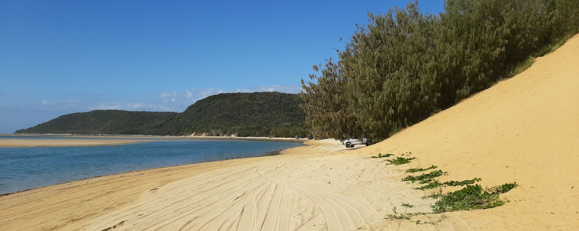 A scenic view of the beach at Double Island Point, Queensland, showcasing the clear turquoise waters, sandy shoreline, and lush mountain backdrop, captured from a 4WD vehicle driving up the beach